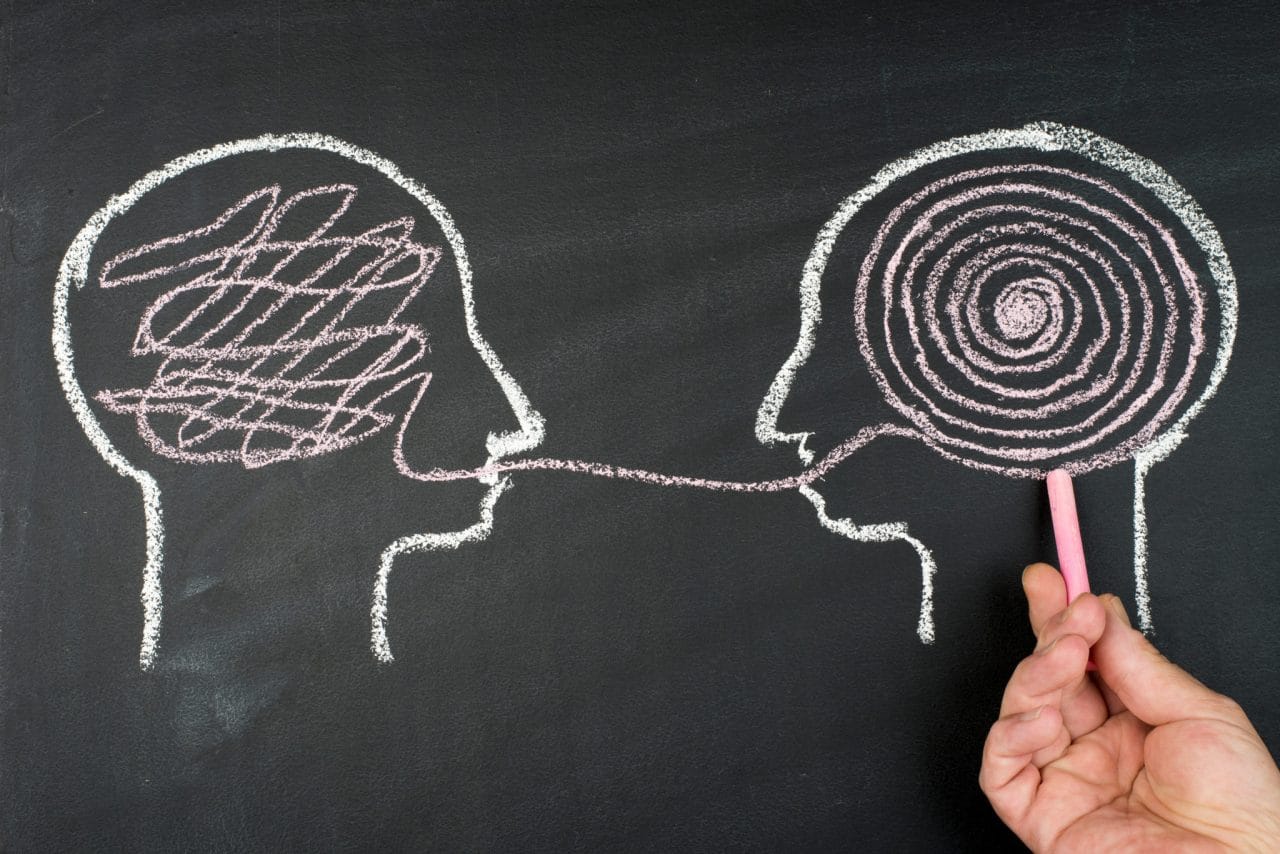 Chalk drawing of two human heads in profile, one's thoughts visualized as a neat spiral while the other's are a tangled scribble. The two are connected through a line that crosses the outline at the mouth of each head.